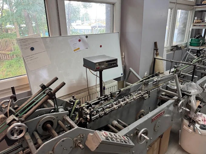 Maszyna introligatorska w warsztacie, widok z przodu. Biała tablica z dokumentami w tle. Sprzęt używany do składania i zszywania arkuszy papieru.