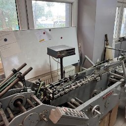 Drukarnia DRUKLANET Przemysław Dąbrowski - Maszyna introligatorska w warsztacie drukarskim, widok z przodu, jasne światło dzienne, biała tablica z notatkami w tle, widoczne elementy mechaniczne.