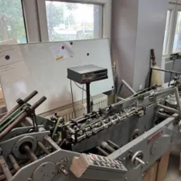 Maszyna introligatorska w warsztacie drukarskim, widok z przodu, jasne światło dzienne, biała tablica z notatkami w tle, widoczne elementy mechaniczne.