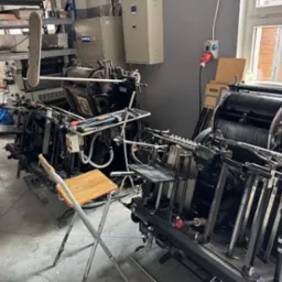 Dwie maszyny drukarskie w hali produkcyjnej, z krzesłem i półkami w tle. Widoczne detale konstrukcyjne maszyn i elementy wyposażenia drukarni.