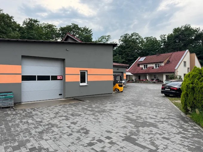Budynek firmy z szarą fasadą i pomarańczowymi pasami, brama garażowa, wózek widłowy, kostka brukowa na podjeździe i dom w tle.