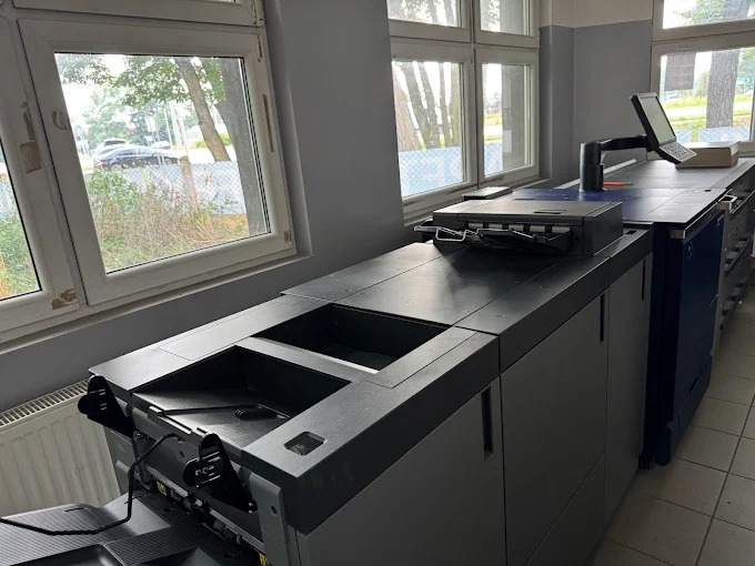 Profesjonalna maszyna drukarska w jasnym pomieszczeniu z oknami, gotowa do druku. Widoczny panel sterowania i podajnik papieru.