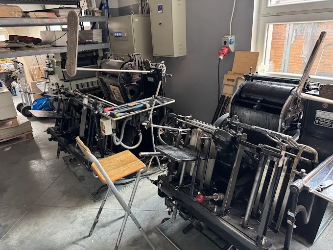 Dwie maszyny drukarskie w hali produkcyjnej, z krzesłem i półkami w tle. Widoczne detale konstrukcyjne maszyn i elementy wyposażenia drukarni.
