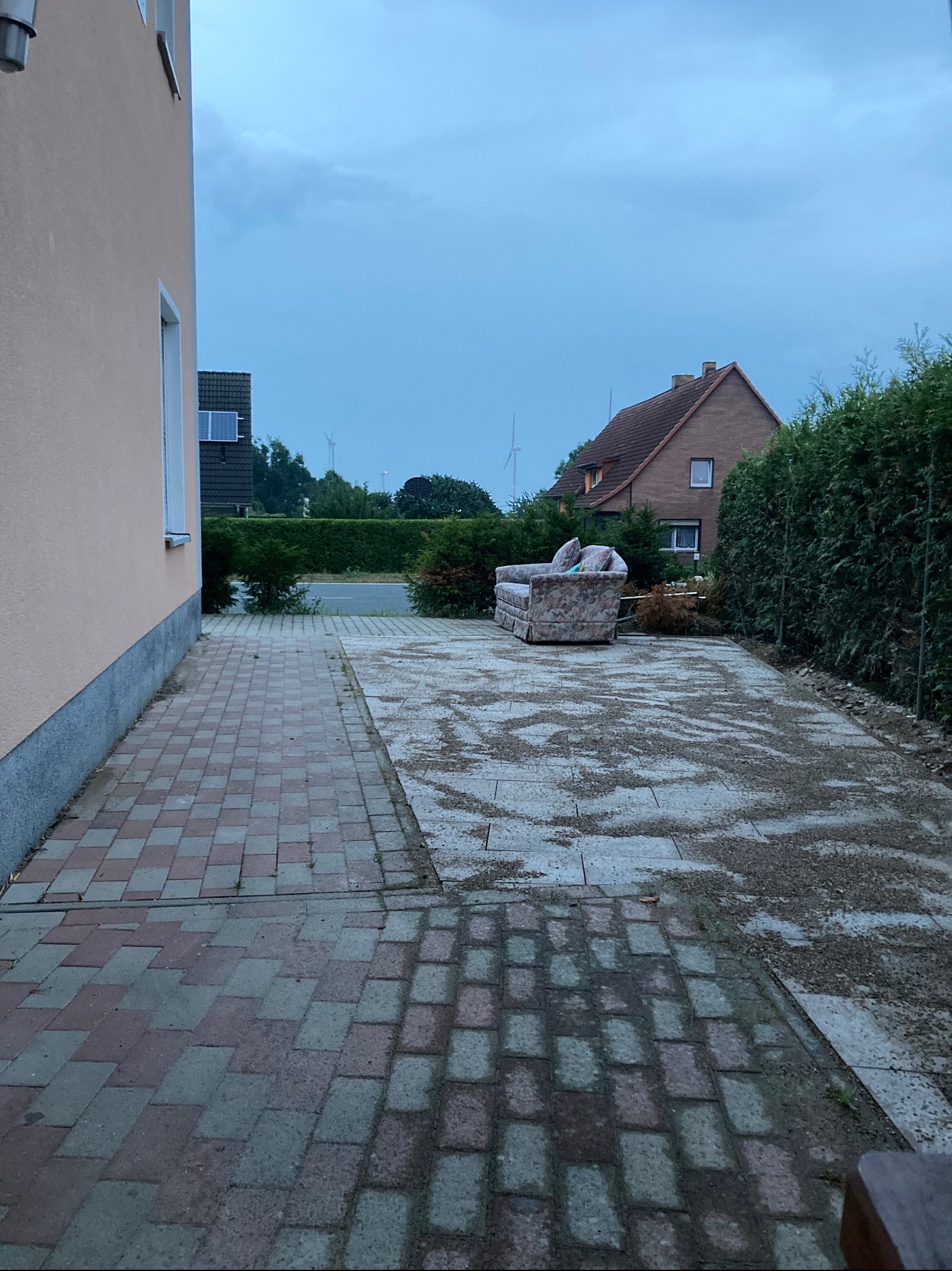 Nierówna nawierzchnia z kostki i gruzu na podwórku z sofą w tle, fragment budynku po lewej, krzewy i dom w oddali, pochmurne niebo.