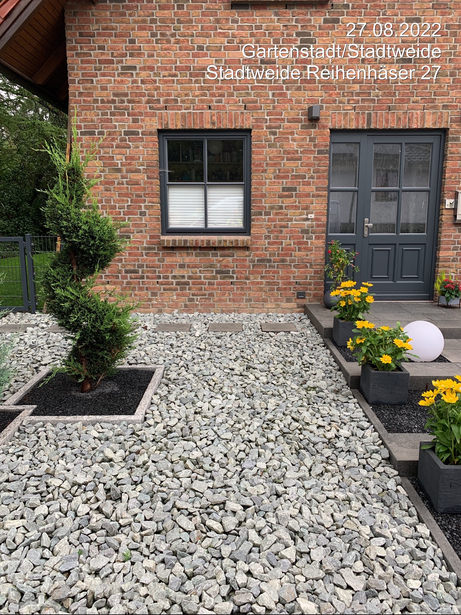 Ogród przed domem z ścieżką z szarego żwiru, ozdobnymi roślinami w donicach i formowaną spiralnie tują w kwadratowej rabacie. Czerwona cegła elewacji.