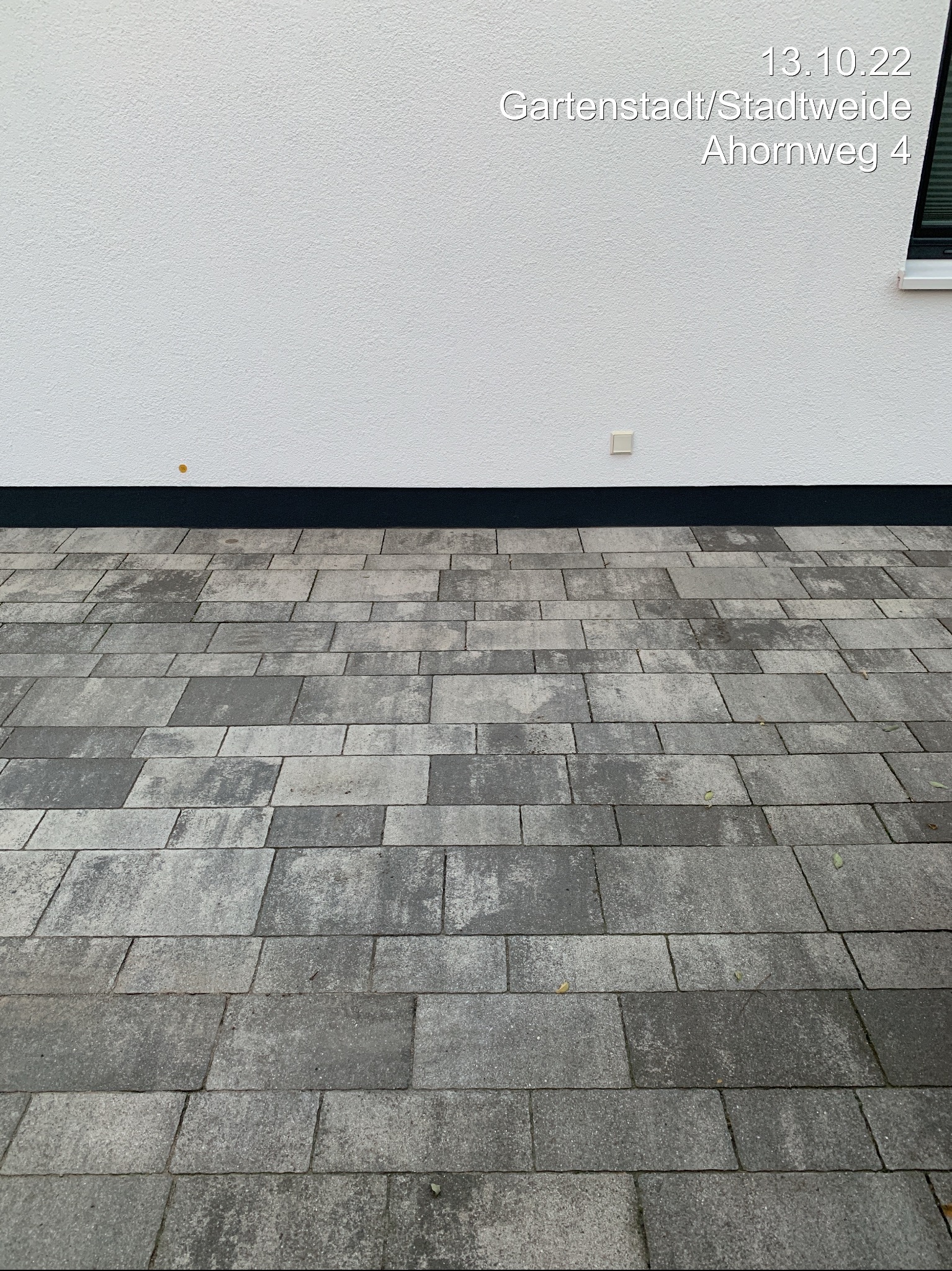 Ułożona szara kostka brukowa na podwórku w Ząbkowicach Śląskich. Widoczna biała ściana budynku z czarnym pasem przy ziemi. Drobne liście na kostce.