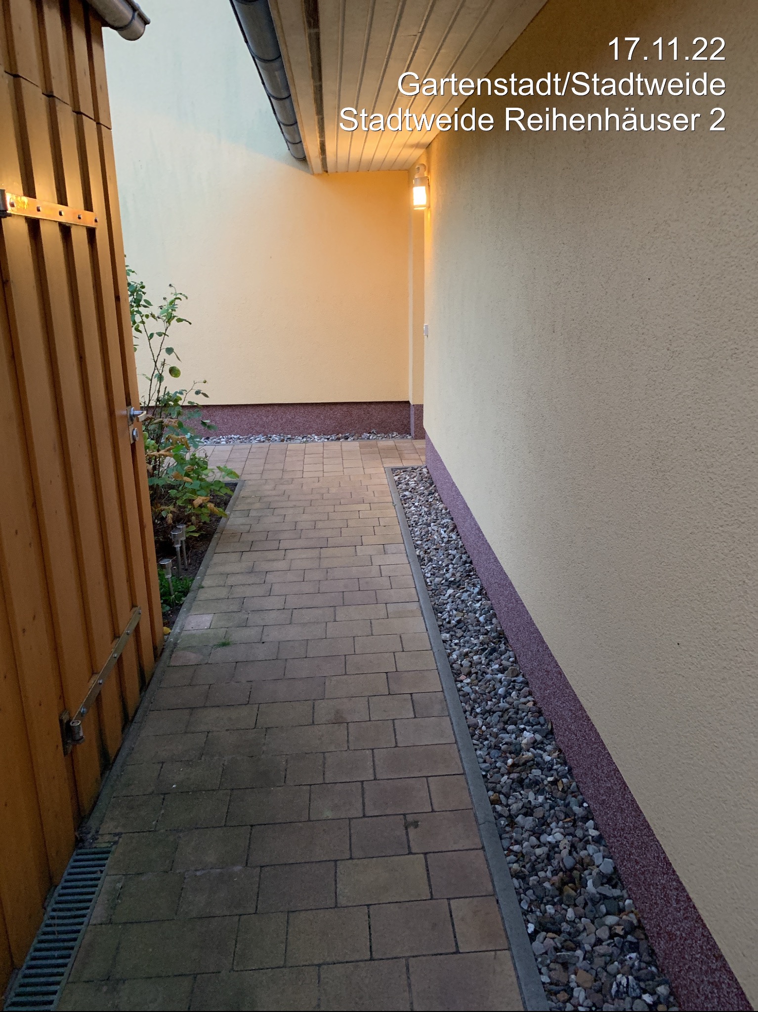 Ułożona kostka brukowa tworzy ścieżkę wzdłuż budynku z żółtą elewacją. Obok ścieżki pas z kamieni, a po lewej drewniany płot. Widoczne oświetlenie zewnętrzne.