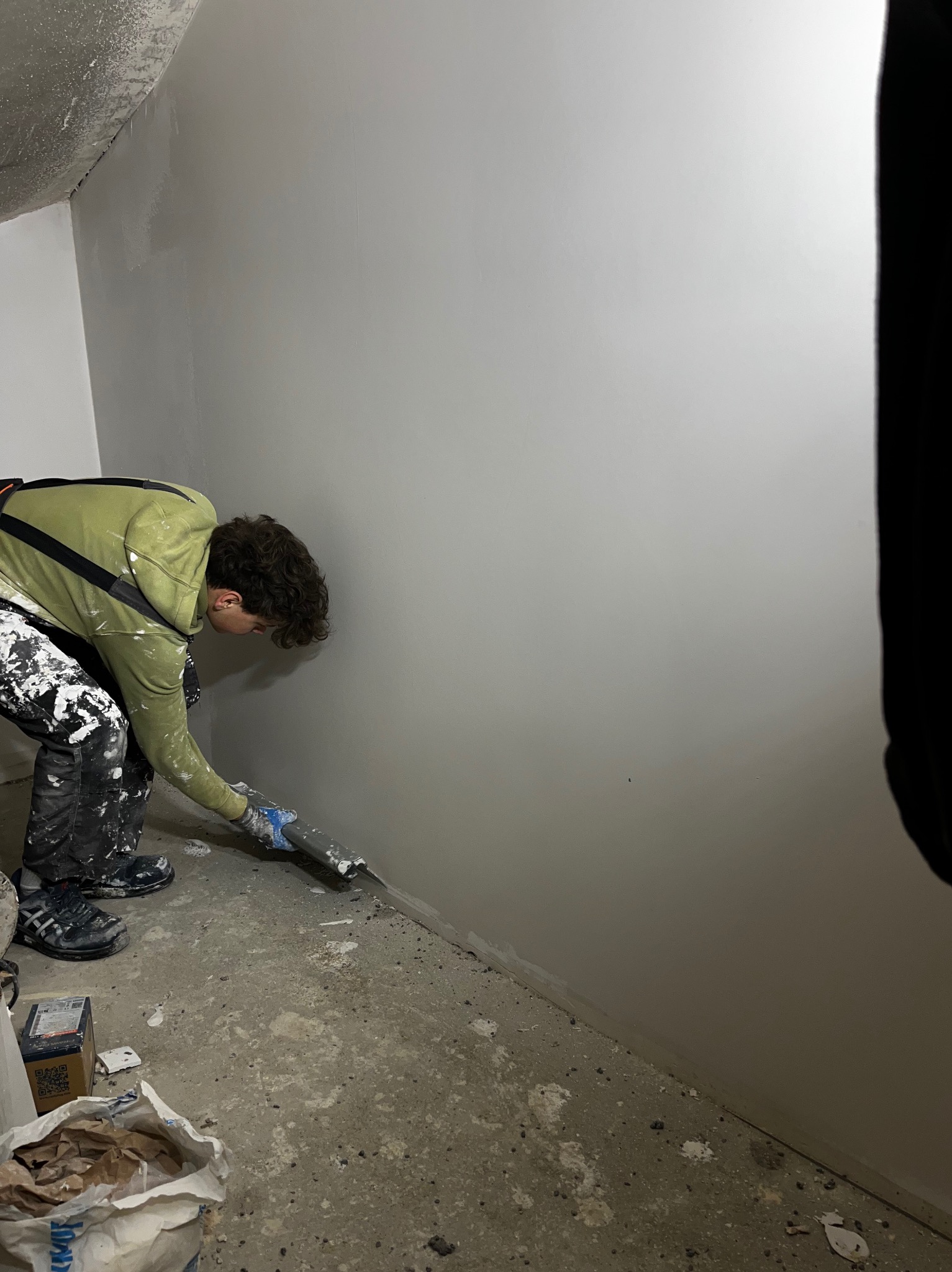 Pracownik w trakcie nakładania gładzi szpachlowej na ścianę w pomieszczeniu. Widoczne narzędzia i materiały budowlane. Ubranie robocze z zabrudzeniami farby.