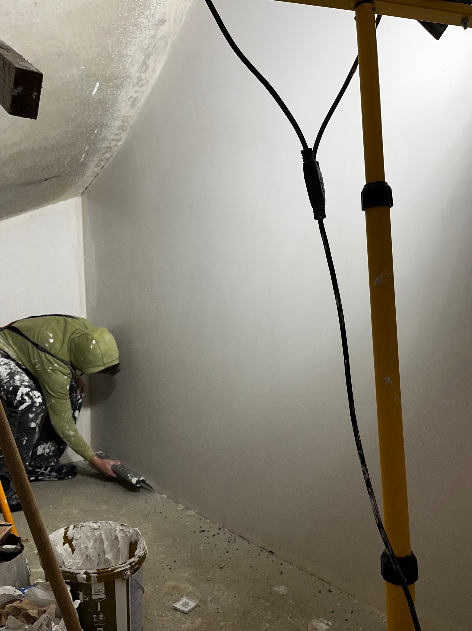 Pracownik w trakcie szpachlowania ściany w pomieszczeniu ze skośnym sufitem, oświetlonym lampą budowlaną. Widoczny kubeł z masą szpachlową i narzędzia.