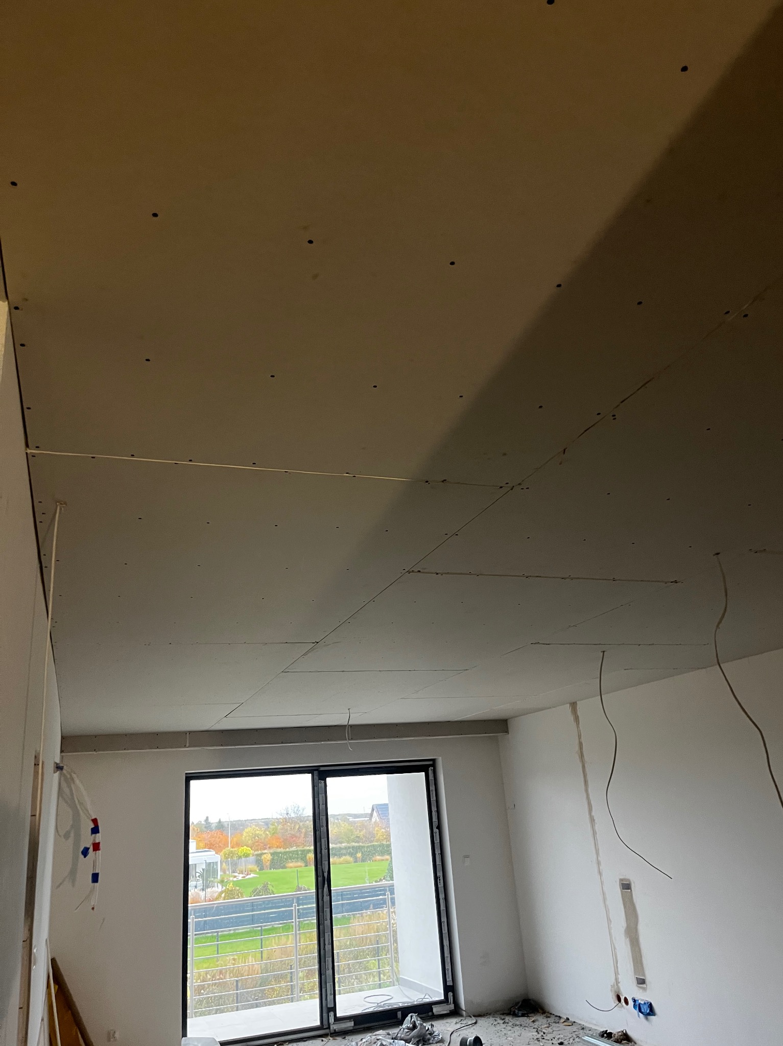 Wnętrze w trakcie prac wykończeniowych: sufit z płyt gipsowo-kartonowych, widoczne przewody elektryczne, okno z widokiem na zieleń. Stan surowy, przygotowanie do dalszych prac.