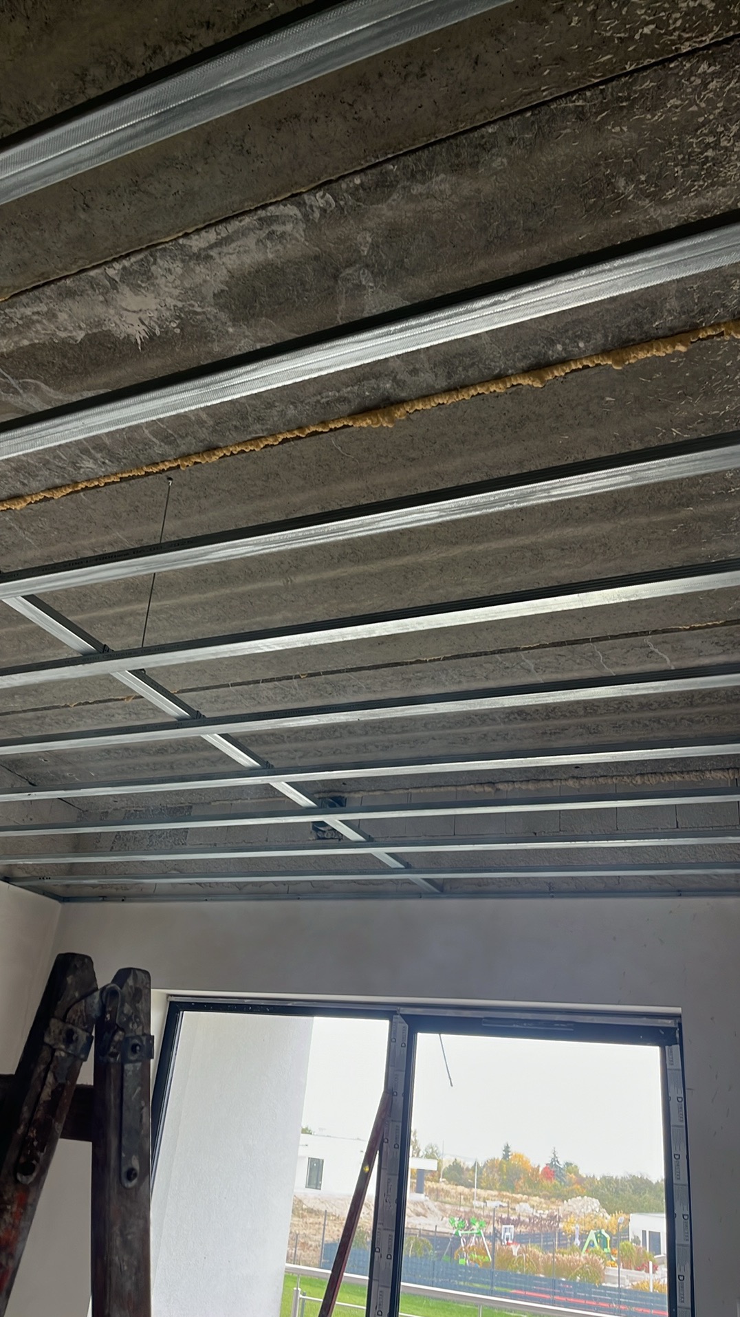 Montaż metalowej konstrukcji sufitu podwieszanego z widoczną pianką montażową, częściowo zasłonięte okno i drabina w tle. Ujęcie z dołu.