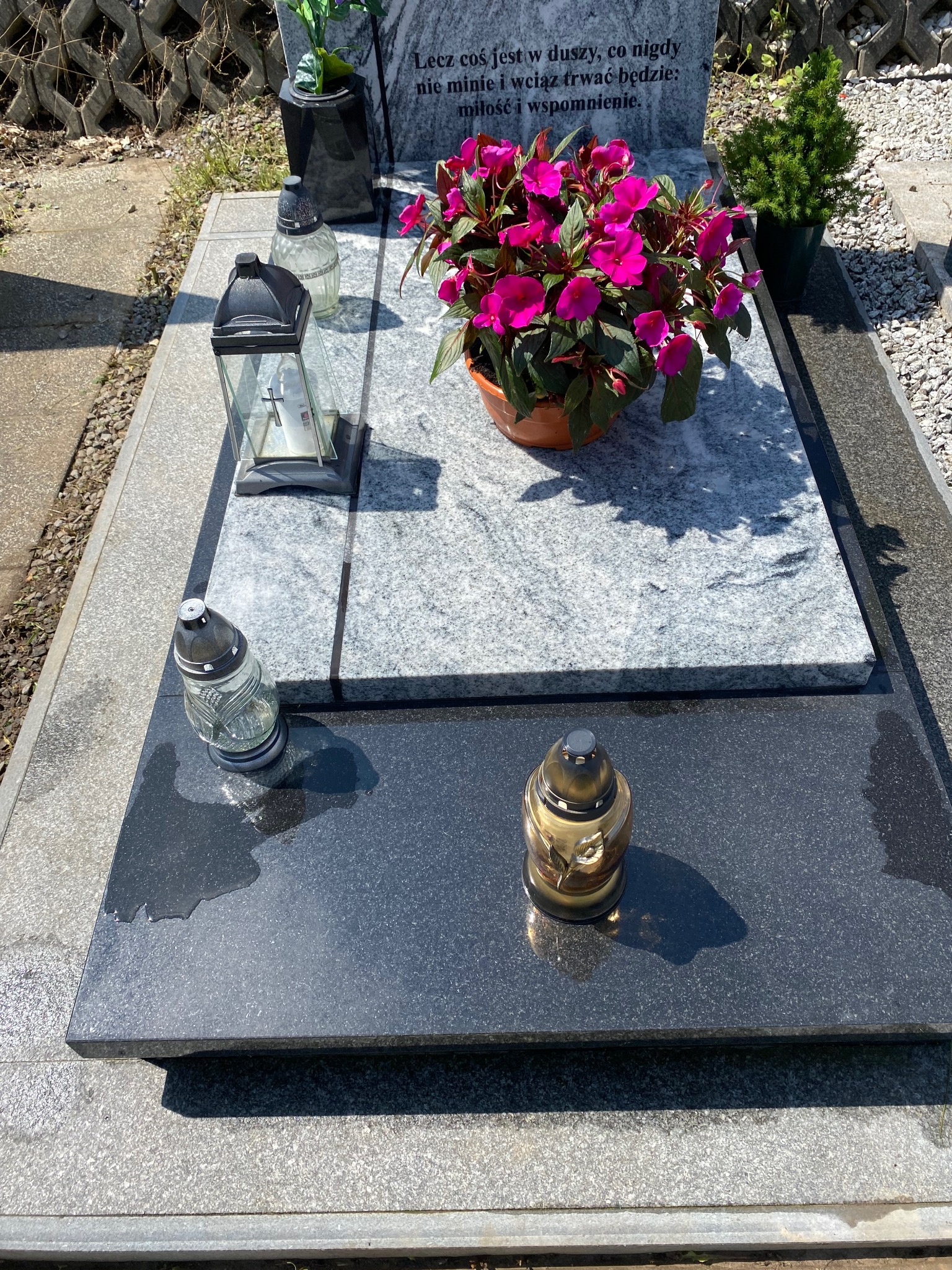 Granitowy nagrobek z kwiatami w doniczce, lampionami i zniczami. Pomnik z wygrawerowanym napisem w słoneczny dzień na cmentarzu.