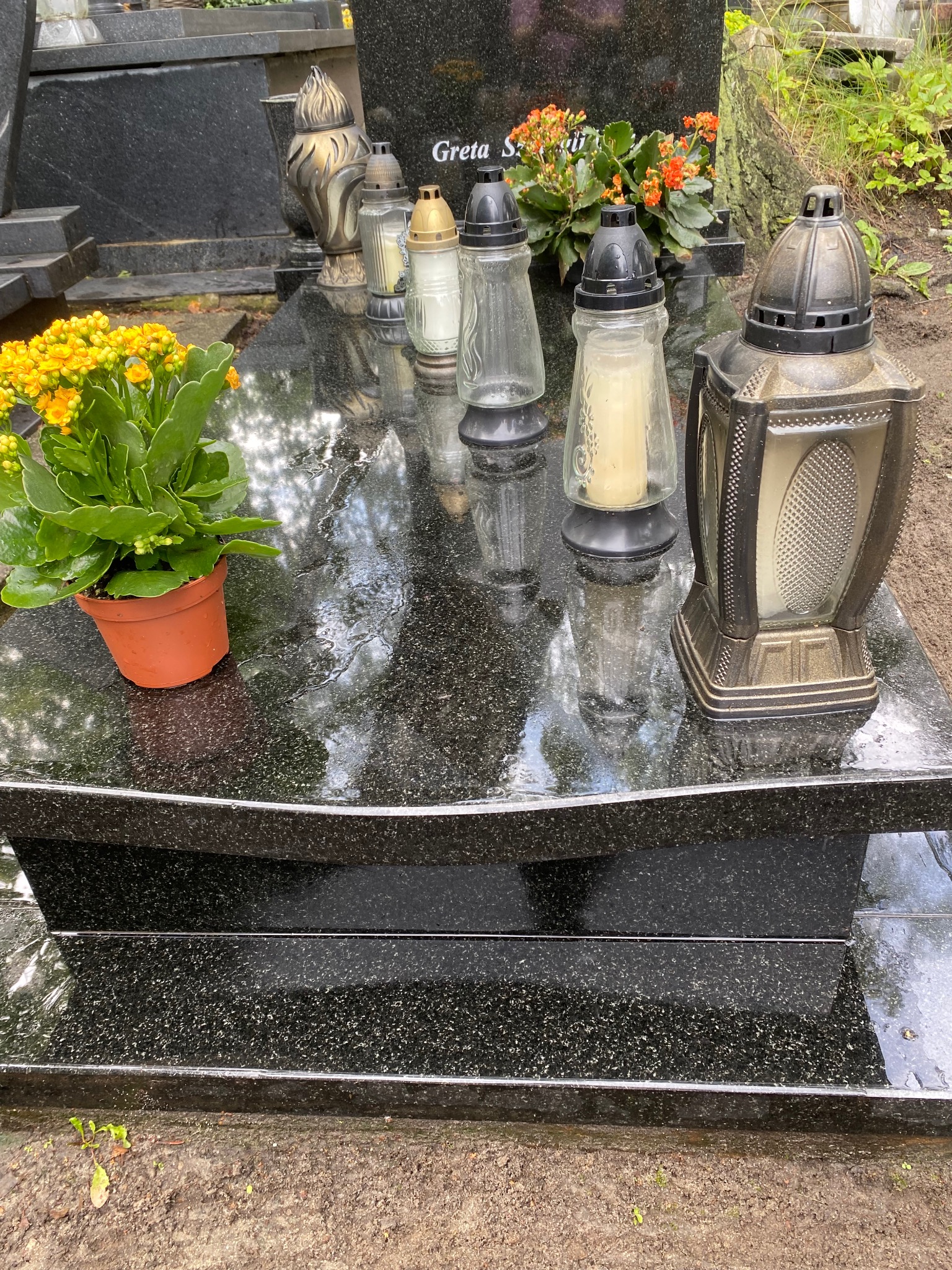 Błyszczący, czarny nagrobek z kwiatami w doniczce i zapalonymi zniczami, po deszczu, z widocznym odbiciem otoczenia.
