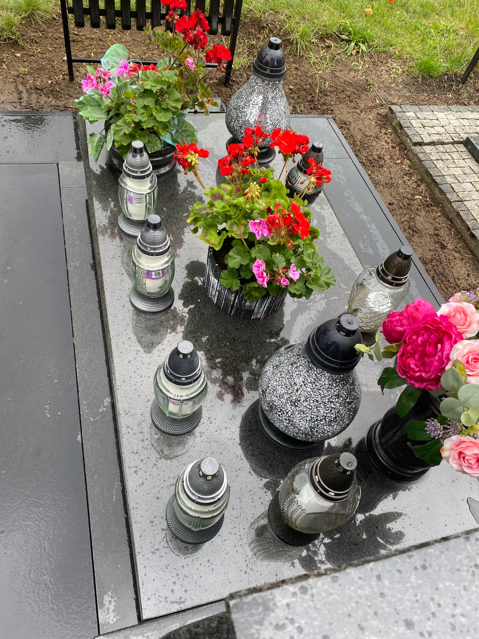 Uporządkowany nagrobek z kwiatami w doniczkach i lampionami, po deszczu. Widoczne ślady wody na czarnym kamieniu nagrobka.