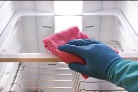 Ręka w niebieskiej rękawicy czyści różową ściereczką wnętrze pustej lodówki z metalowymi półkami. Widok z góry, oświetlenie górne.