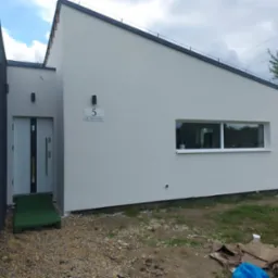 Elewacja nowoczesnego domu jednorodzinnego z numerem '5', szara fasada, okno z białymi ramami, drzwi wejściowe z rampą, widok z zewnątrz w słoneczny dzień.