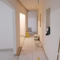 Paweł Tomalski - Korytarz w trakcie układania białych płytek z marmurowym wzorem, widoczne narzędzia: poziomica, worki z klejem, kable, listwy. Ściany białe, sufit z oświetleniem.