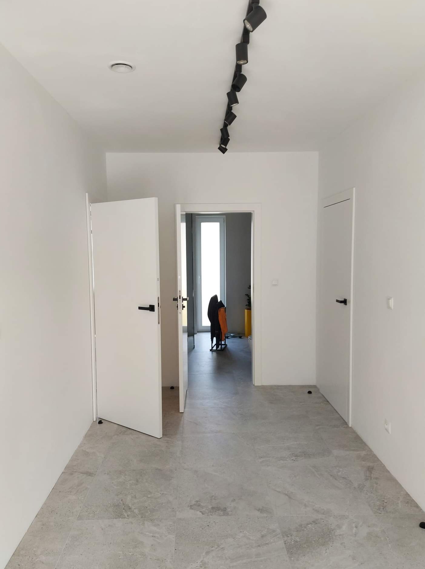 Jasne wnętrze korytarza po remoncie z szarą podłogą z płytek, białymi ścianami i dwiema parami białych drzwi z czarnymi klamkami. Oświetlenie sufitowe punktowe.