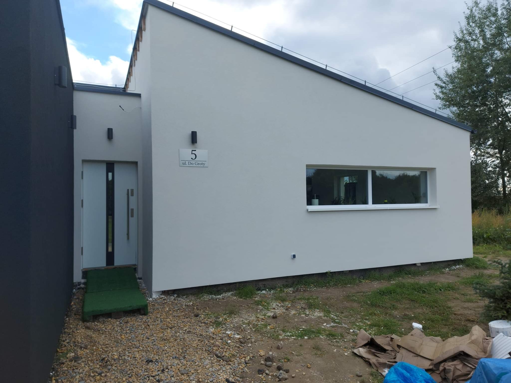 Elewacja nowoczesnego domu jednorodzinnego z numerem '5', szara fasada, okno z białymi ramami, drzwi wejściowe z rampą, widok z zewnątrz w słoneczny dzień.