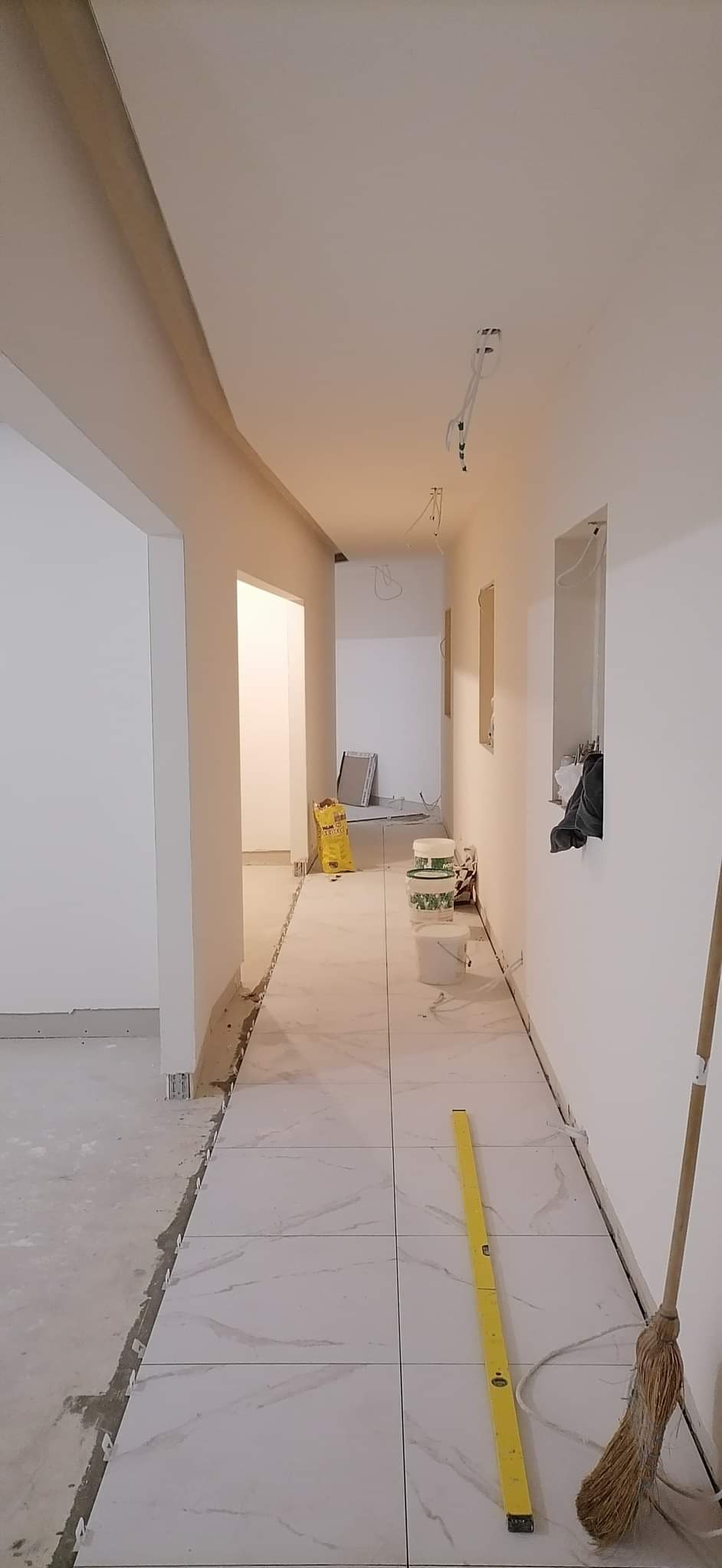 Korytarz w trakcie układania białych płytek z marmurowym wzorem, widoczne narzędzia: poziomica, worki z klejem, kable, listwy. Ściany białe, sufit z oświetleniem.