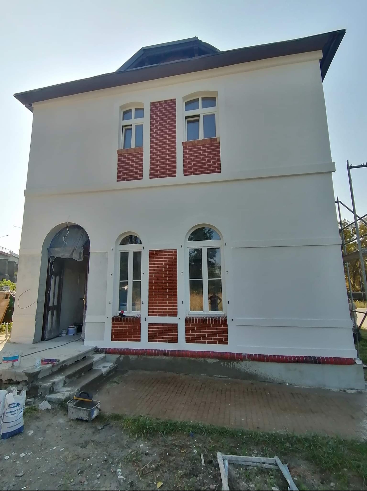 Dwukondygnacyjny budynek po renowacji elewacji: białe ściany z ceglanymi akcentami wokół okien i u podstawy. Widoczne materiały budowlane i narzędzia.