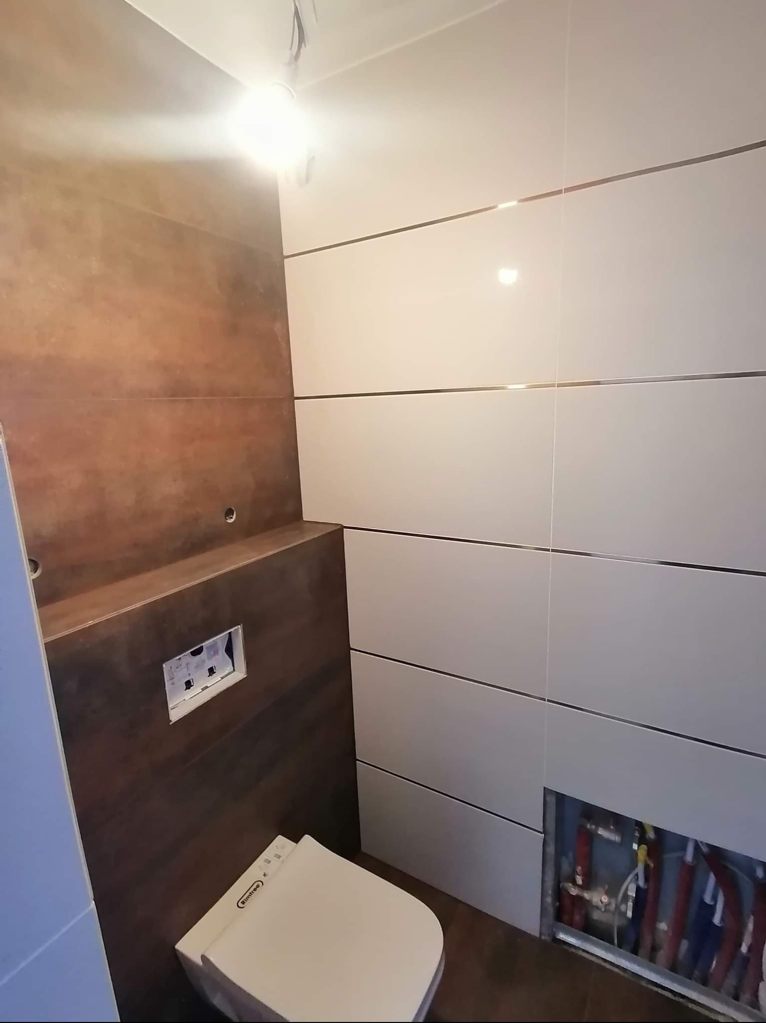 Nowoczesna łazienka w trakcie wykańczania: białe i brązowe płytki z metalowymi listwami, częściowo widoczna instalacja sanitarna i podwieszana toaleta.