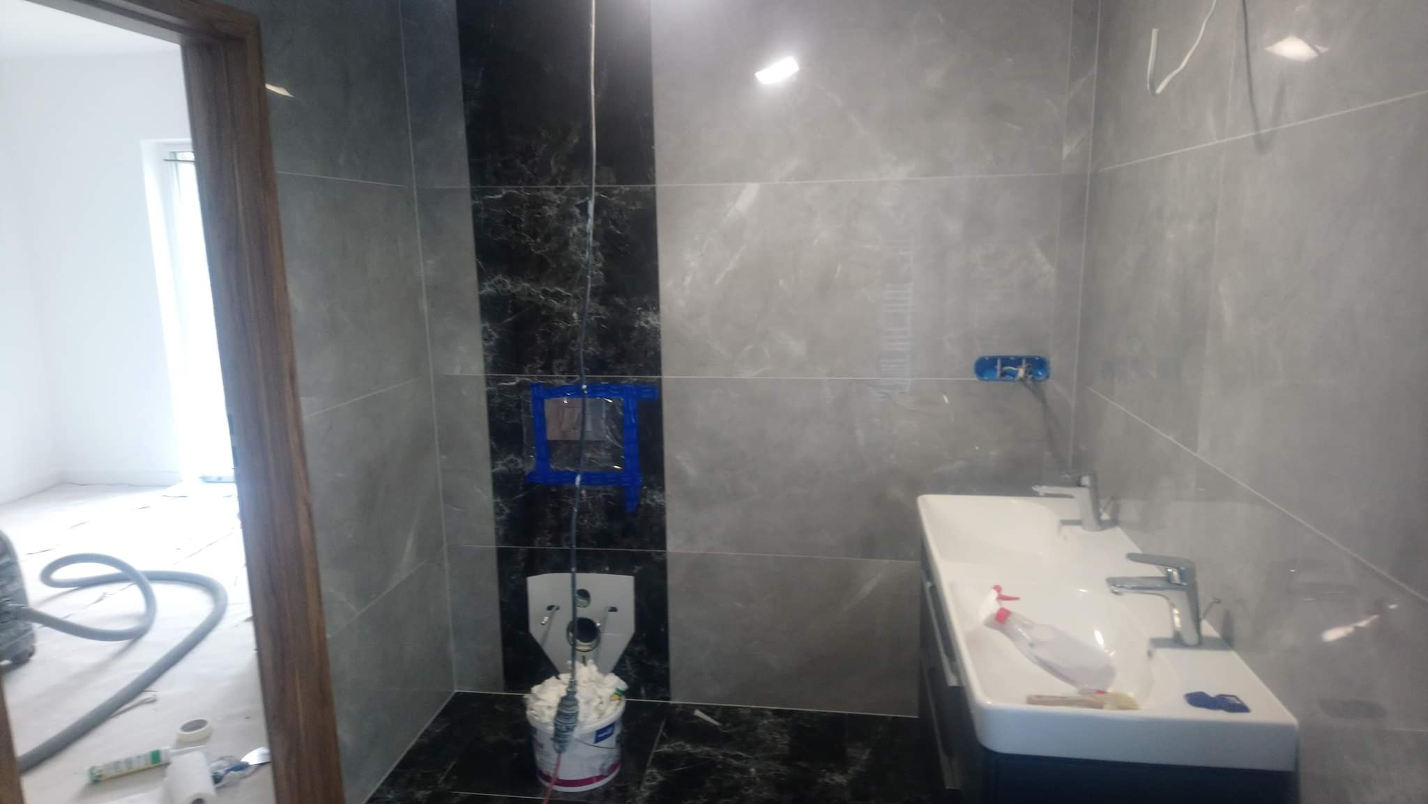 Łazienka w trakcie wykańczania: szare płytki imitujące marmur, czarny pas z wzorem marmuru, biała umywalka i bateria. Widoczne przygotowania do montażu toalety.