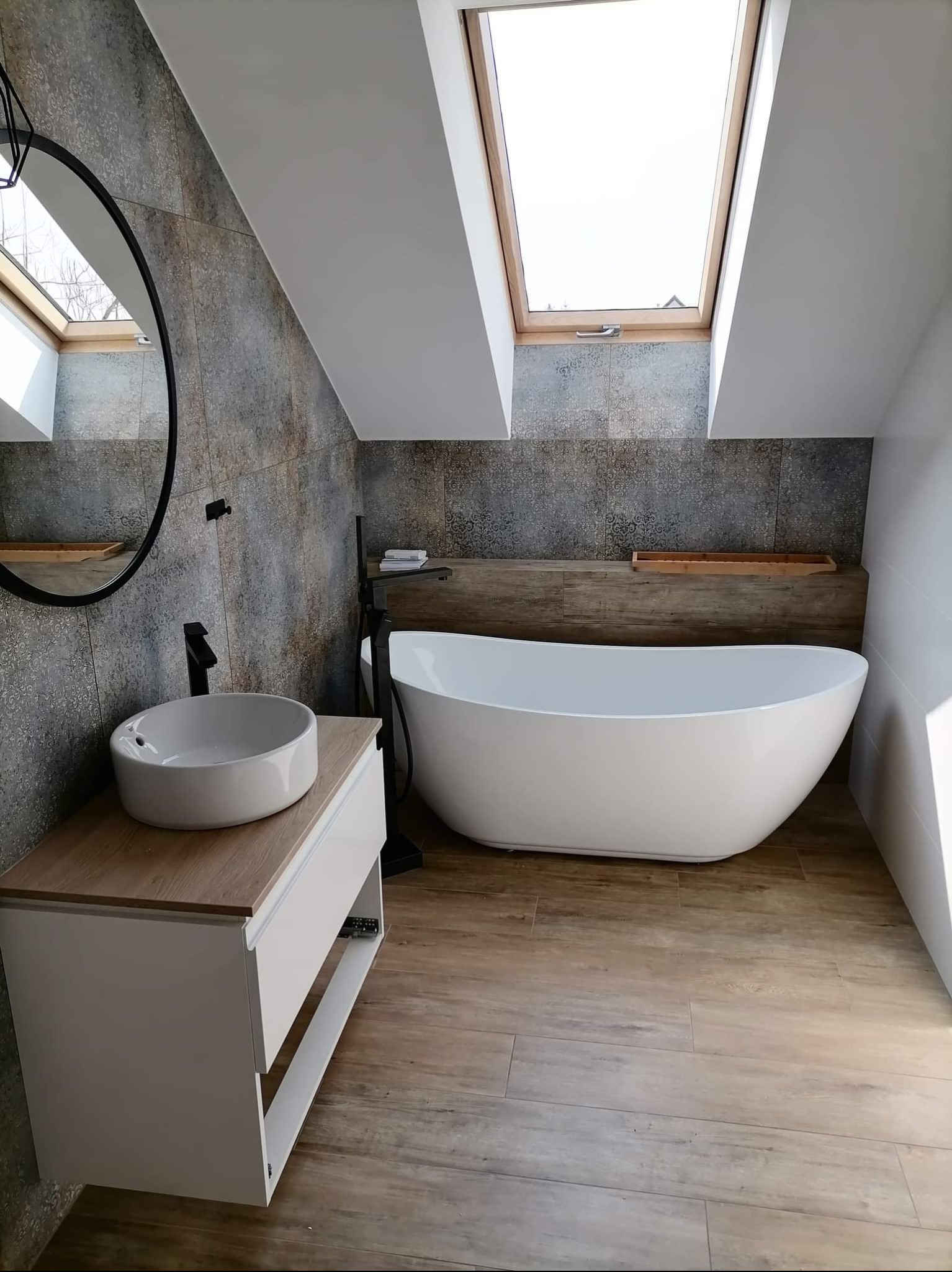 Nowoczesna łazienka na poddaszu z wanną wolnostojącą, umywalką nablatową i oknem dachowym. Płytki imitujące drewno na podłodze i dekoracyjne na ścianach.