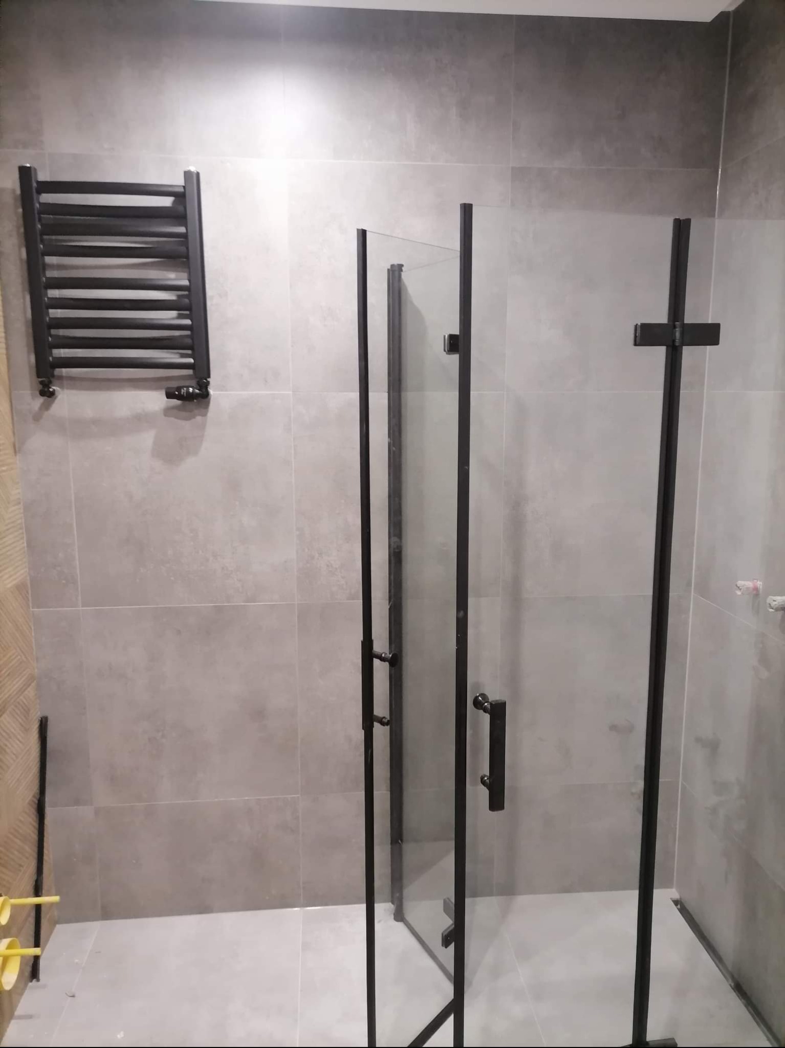 Nowoczesna łazienka po remoncie: szare płytki, czarna kabina prysznicowa typu walk-in i grzejnik drabinkowy. Minimalistyczny design.
