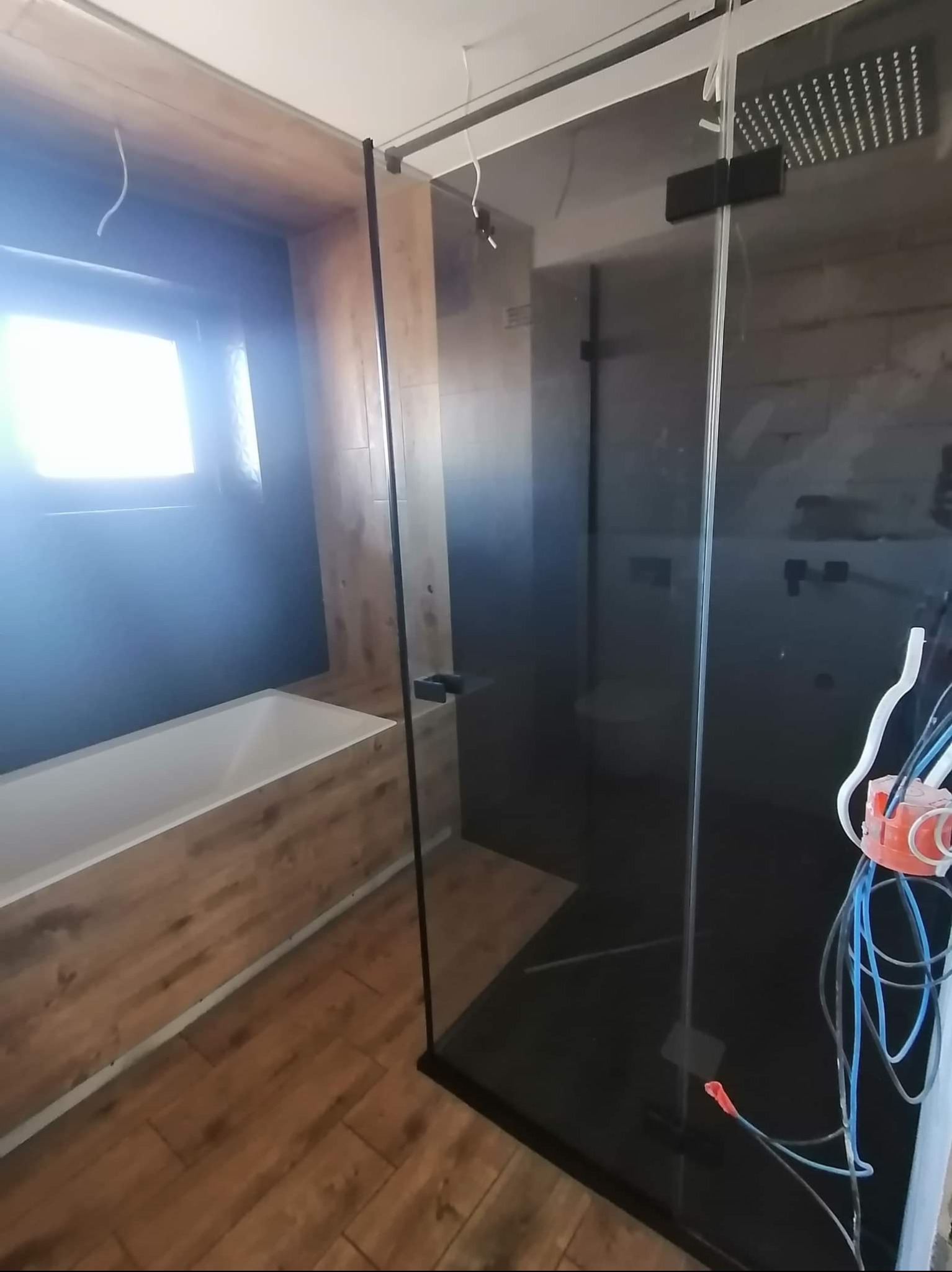 Nowoczesna łazienka z kabiną typu walk-in, wykończona drewnopodobnymi płytkami. Widoczna wanna obudowana drewnem i czarna ściana. Prace wykończeniowe w toku.
