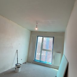 JC Interiors - Pokój po szpachlowaniu ścian i sufitu, widoczne narzędzia, okno zabezpieczone folią, gotowe do malowania. Ściany gładkie, jasne, surowe oświetlenie.