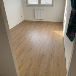 JC Interiors - Jasne panele podłogowe ułożone w pustym pokoju z białymi ścianami i oknem. Widoczny fragment futryny drzwi.