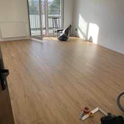 JC Interiors - Jasne panele podłogowe w pokoju z białymi ścianami i otwartym balkonem. Widoczne narzędzia i akcesoria do montażu podłogi. Naturalne światło słoneczne.