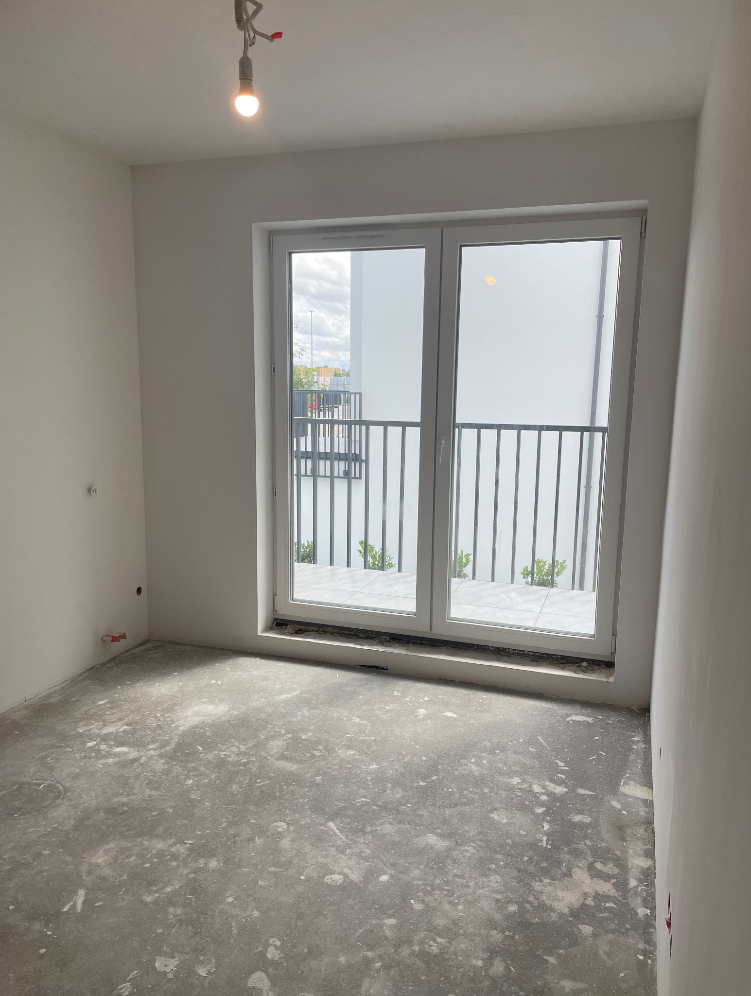 Surowe wnętrze z dużym oknem balkonowym i wylewką na podłodze, przygotowane do dalszych prac wykończeniowych. Widoczna żarówka.