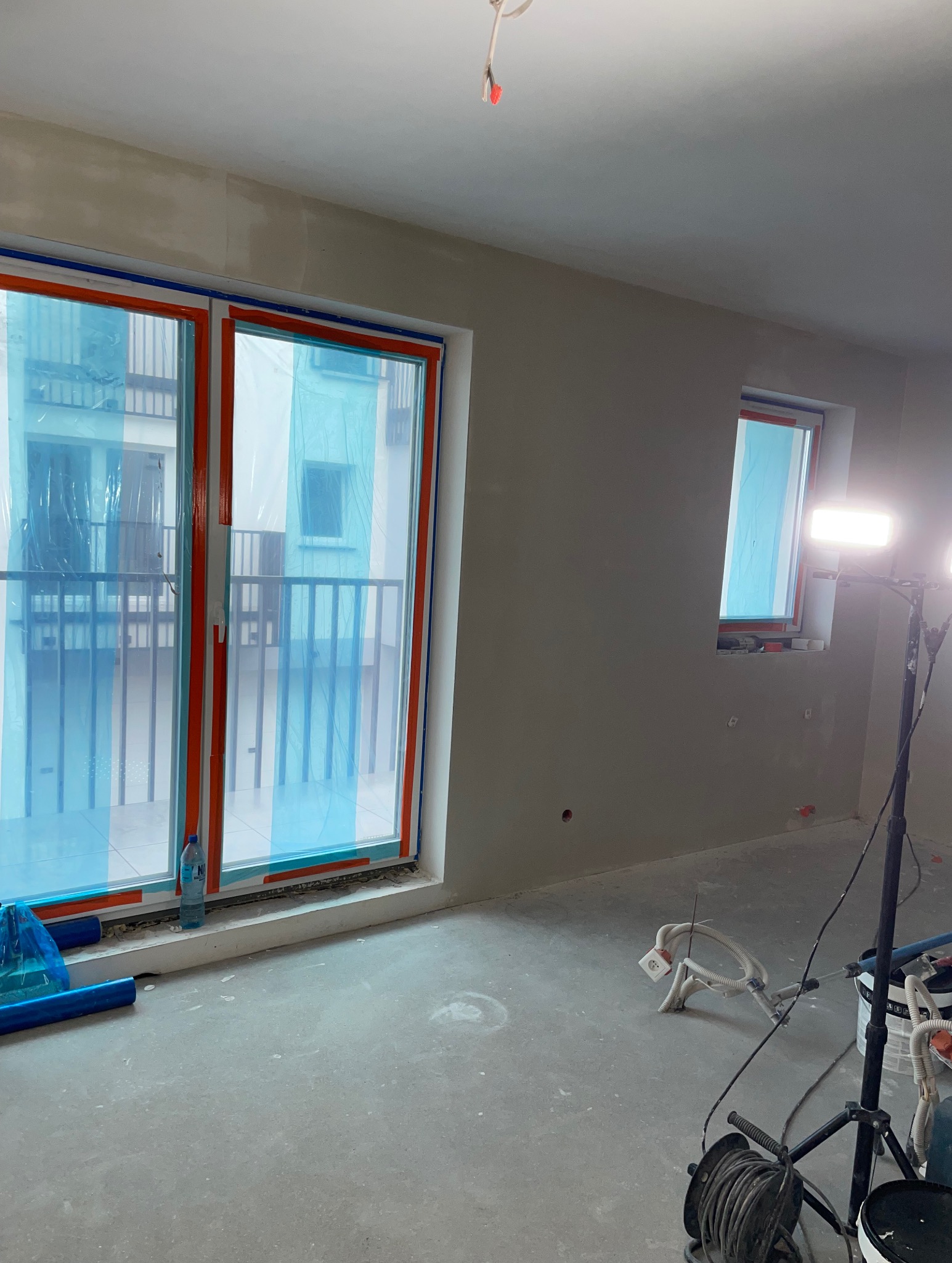 Pokój po szpachlowaniu ścian, przygotowany do malowania. Okna zabezpieczone folią, widoczne narzędzia i oświetlenie budowlane. Szare ściany i podłoga.
