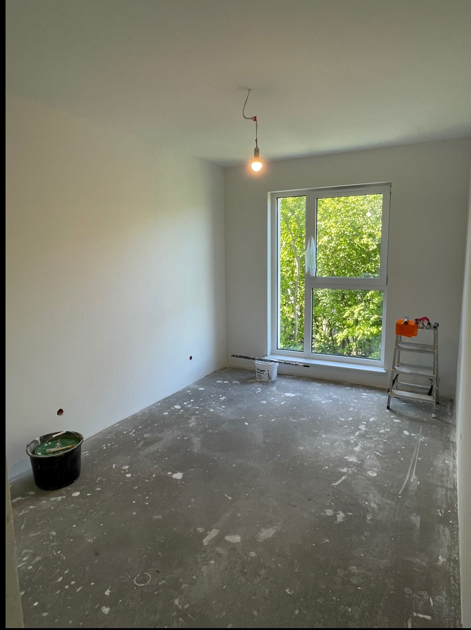 Pomalowane na biało wnętrze pokoju z widoczną drabiną, wiadrem z farbą i oknem z zielonym widokiem. Podłoga z plamami farby. Żarówka zwisa z sufitu.
