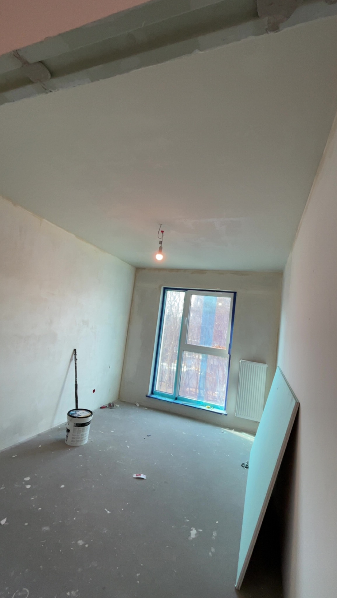 Pokój po szpachlowaniu ścian i sufitu, widoczne narzędzia, okno zabezpieczone folią, gotowe do malowania. Ściany gładkie, jasne, surowe oświetlenie.