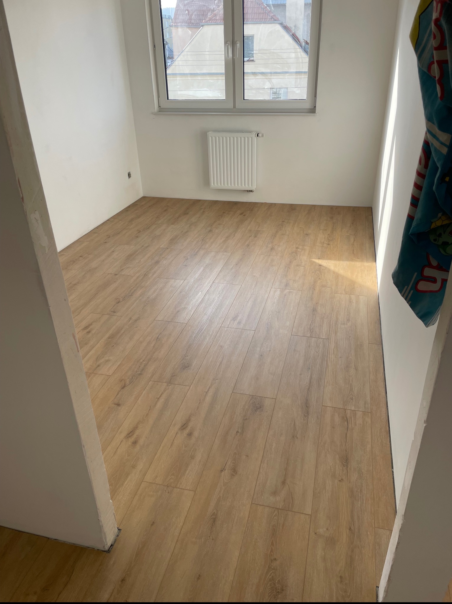 Jasne panele podłogowe ułożone w pustym pokoju z białymi ścianami i oknem. Widoczny fragment futryny drzwi.