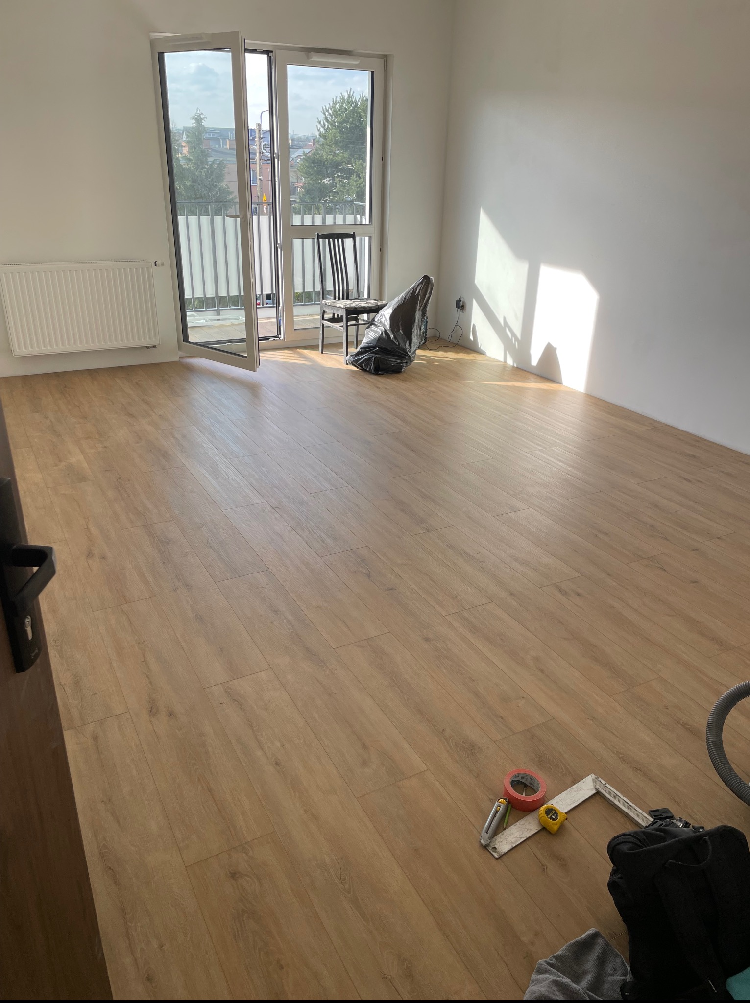 Jasne panele podłogowe w pokoju z białymi ścianami i otwartym balkonem. Widoczne narzędzia i akcesoria do montażu podłogi. Naturalne światło słoneczne.