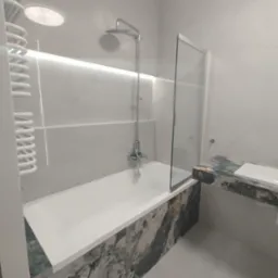 Elegancka łazienka w jasnych odcieniach szarości, z wanną obudowaną płytkami imitującymi marmur i szklanym parawanem. Oświetlenie LED podkreśla nowoczesny charakter.