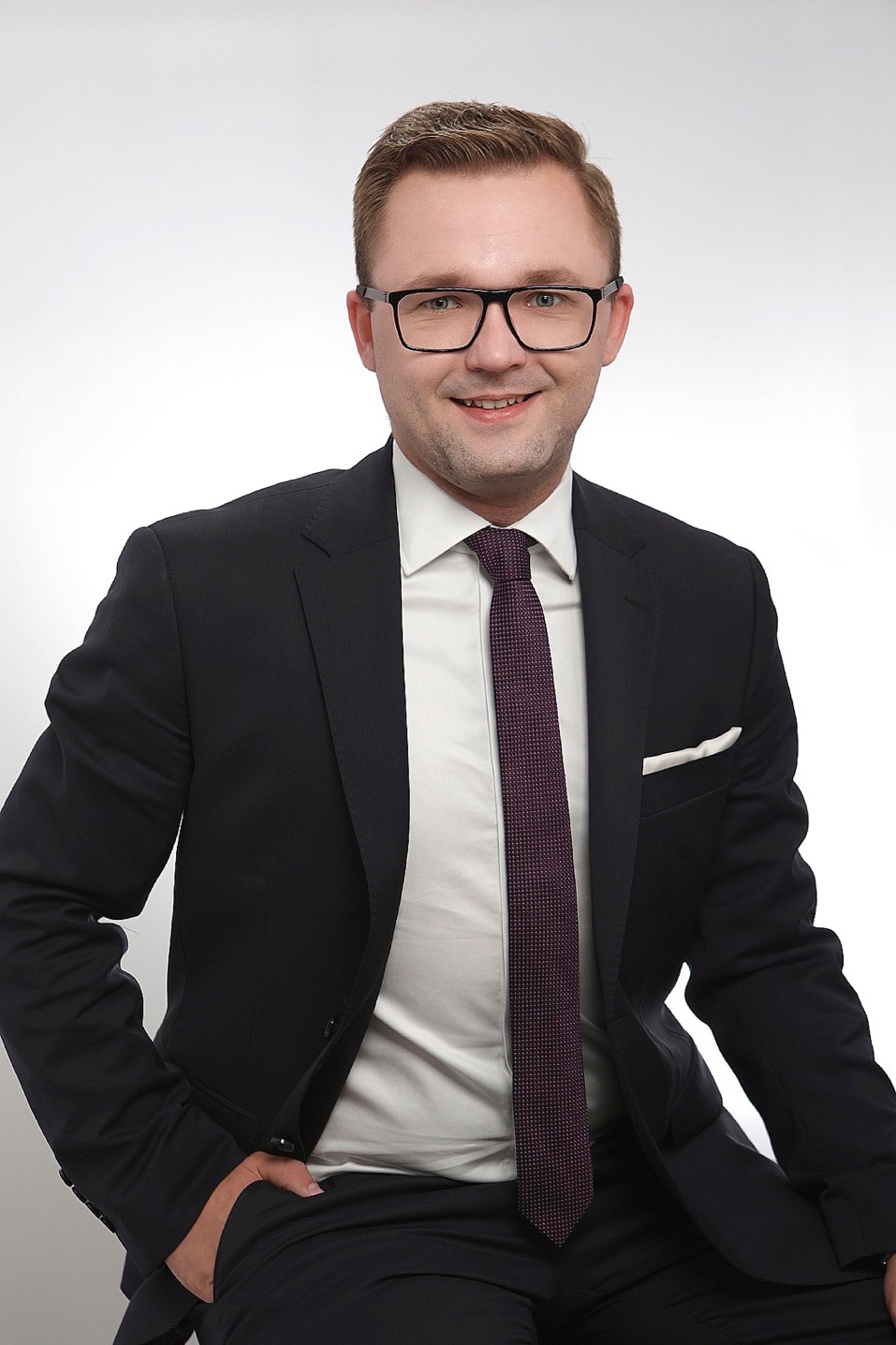 Portret mężczyzny w okularach, ciemnym garniturze i bordowym krawacie, z białą poszetką w kieszonce, na jasnym tle. Uśmiechnięty, profesjonalny wizerunek.