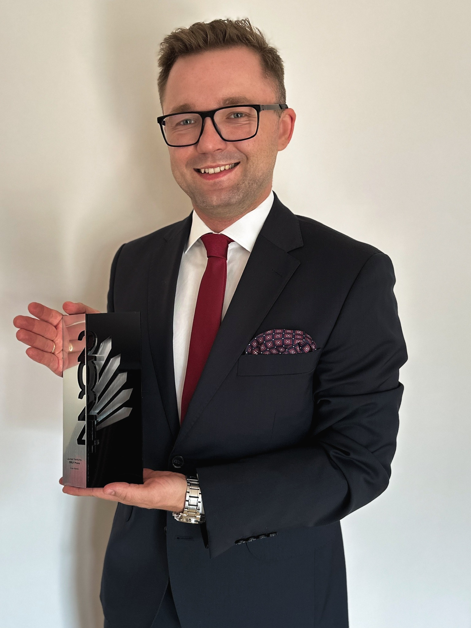 Uśmiechnięty mężczyzna w garniturze prezentuje nagrodę 'Laureat Konkursu O!PLY Prawa' na tle białej ściany. Widoczny zegarek i poszetka w kieszonce.