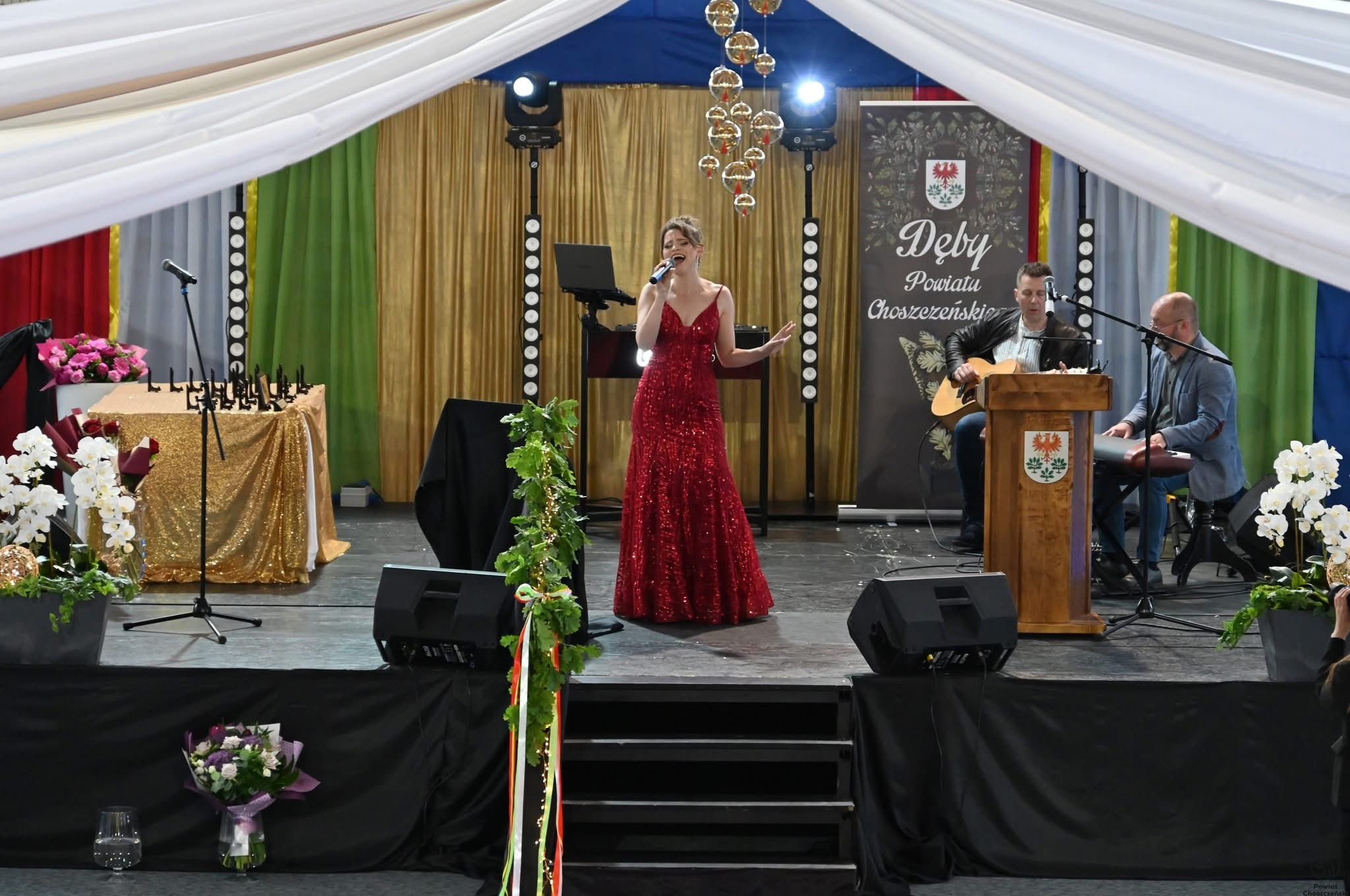Wokalistka w czerwonej sukni śpiewa na scenie podczas gali, akompaniują jej gitarzysta i klawiszowiec. Scena udekorowana jest kwiatami i oświetleniem.