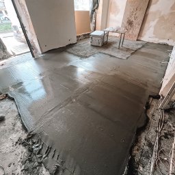 Yauheni Zabarouski - Wylewka podłogowa samopoziomująca w trakcie prac. Widoczne kable elektryczne, płyty drewniane i okno. Nierówności podłoża częściowo zakryte wylewką.