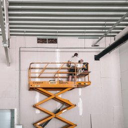 Yauheni Zabarouski - Praca na podnośniku: montaż płyt gipsowych na ścianie w surowym wnętrzu. Widoczne instalacje, oświetlenie sufitowe i fragment otwartego pomieszczenia w tle.