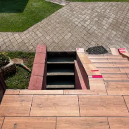 Wykończenie schodów zewnętrznych płytkami imitującymi drewno, prowadzących do piwnicy. Widoczne fragmenty trawnika i kostki brukowej. Czerwone akcenty na krawędziach schodów.