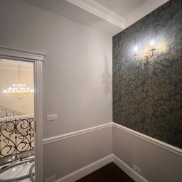 Yauheni Zabarouski - Eleganckie wykończenie wnętrza: ściana z wzorzystą tapetą i kinkietem, listwy przypodłogowe, widok na schody z kryształowym żyrandolem przez otwarte drzwi.