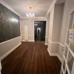 Yauheni Zabarouski - Korytarz po remoncie z ciemną drewnianą podłogą, szarą tapetą w kwiaty i kryształowym żyrandolem. Widoczne listwy przypodłogowe i drzwi z napisem.