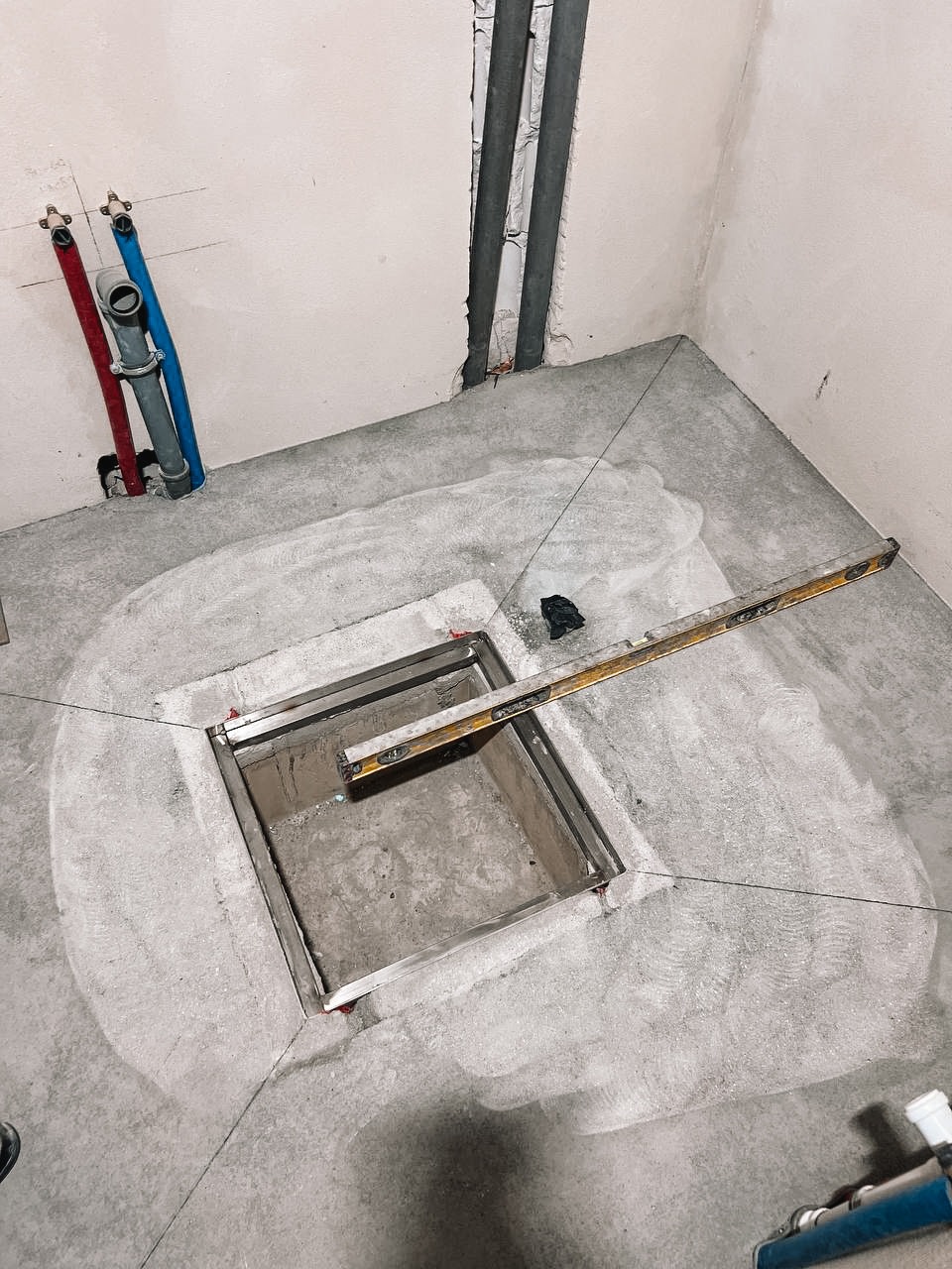 Nierówna, betonowa podłoga z wbudowanym odpływem liniowym w łazience w trakcie prac wykończeniowych. Widoczne rury i poziomica.