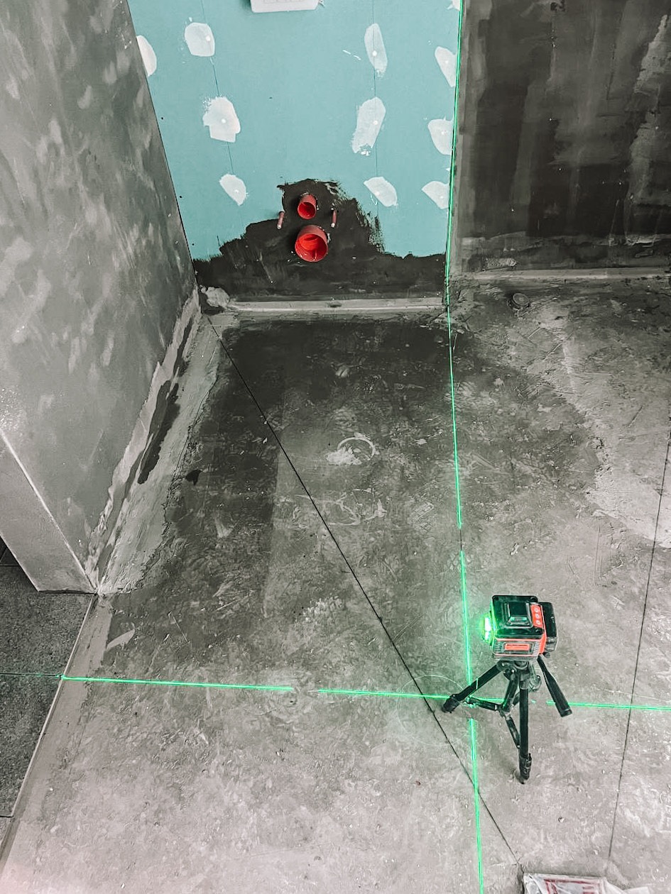 Surowa łazienka w trakcie prac wykończeniowych: ściany przygotowane, widoczne rury i poziomica laserowa z zielonymi liniami.